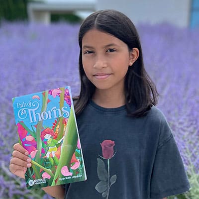 A girl holding a "Path of Thorns" book outdoors in a lavender field, promoting literacy and youth empowerment through Ripple Foundation programs.